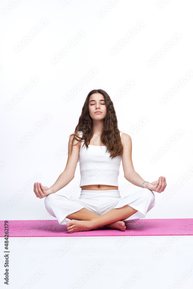 Teenage girl practicing meditation with her eyes closed, sitting in lotus position, on a pink yoga mat Teenage girl practicing meditation with her eyes closed, sitting in lotus position, on a pink yoga mat