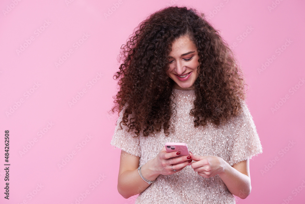 A curly-haired woman typing a message on her smart phone, studio pink background A curly-haired woman typing a message on her smart phone, studio pink background