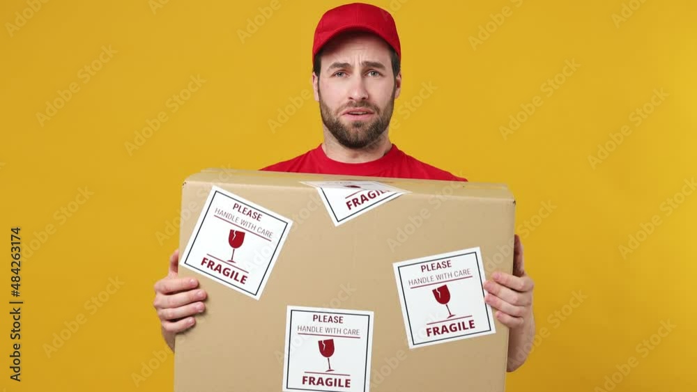 Anxious professional delivery employee man in red cap T-shirt uniform work as dealer courier hold give fragile cardboard box you isolated on plain yellow background studio portrait. Service concept