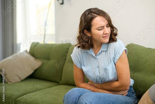 Young caucasian woman with curly hairstyle sitting at the sofa and suffering from stomach ache, keeping hands on her stomach at home, waiting for doctor, poisoning, menstruation