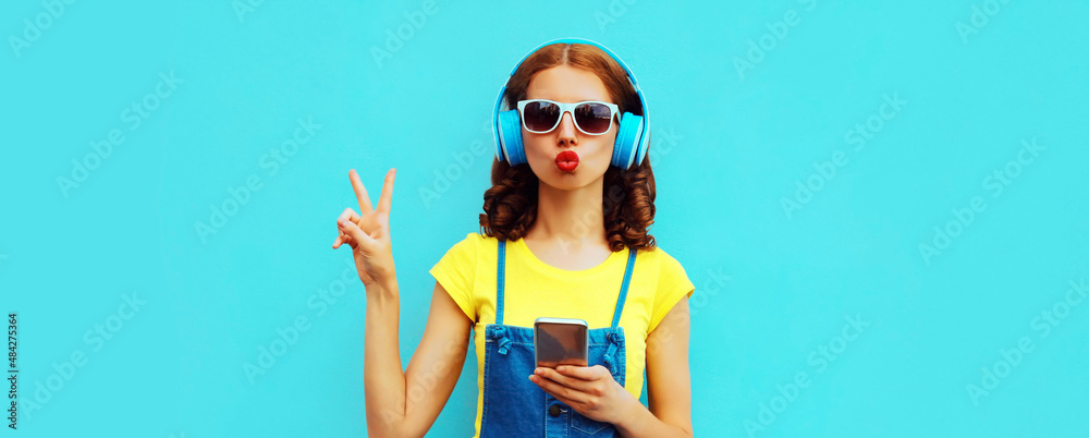 Fototapeta premium Portrait of young woman in headphones listening to music with phone on blue background