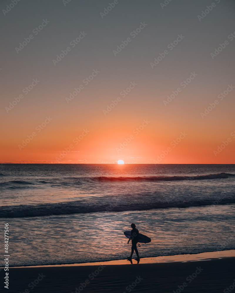 Silhouette of a person with a surf board walking by the beach at sunset