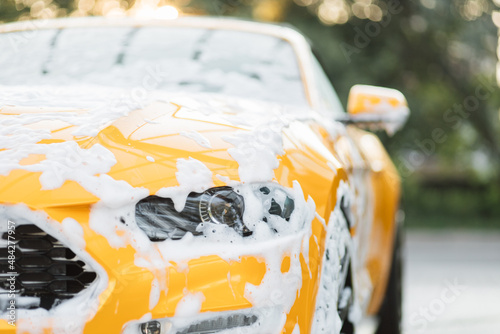 Close up horizontal shot of yellow car headlight with cleaning foam, washed at car wash service outdoors with high pressure sprayer. Clean car concept. Car wash outdoor. photo