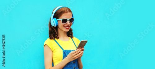 Portrait of happy smiling young woman in headphones listening to music with phone on blue background