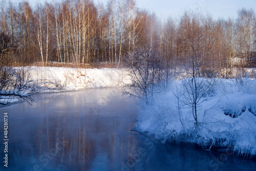 Winter landscape in the Moscow region. The Pekhorka River with non-freezing water. Snow-covered shores, steam rising from the water.  © Svetlana