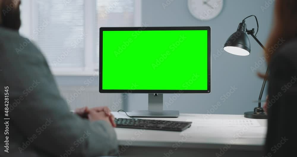 Back view of male and female business colleagues sitting together at office desk and looking on computer with green blank screen. People, cooperation and work concept.