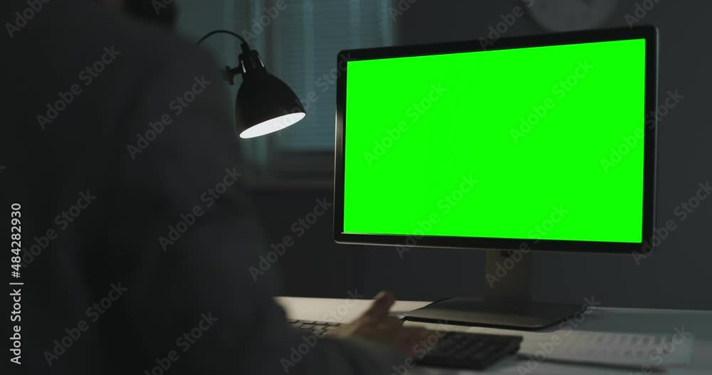 Back view of business person using modern pc while working late at dark office. Focus on chroma key computer screen. Overwork concept.