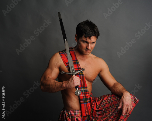 The sexy highlander is posing indoor wearing a medieval costume and holding a sword.  photo