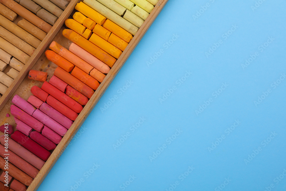 Set of soft pastels in wooden box on light blue background, top view with space for text. Drawing material