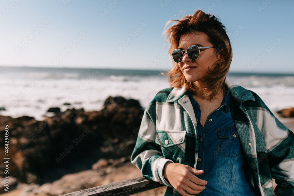 Stylish adorable girl with short blowing hair in sunglasses and striped shirt looking aside and smile on nature background. Outdoor photo of carefree european lady chilling near the sea.