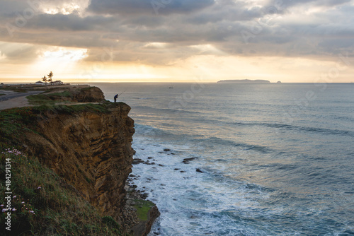 Sunset landscape with a silhouette of a fisherman in the sun on the shores of the Atlantic Ocean on the edge of a sheer high cliff and foamy waves. A small island is visible on the horizon. Peniche © Ekaterina