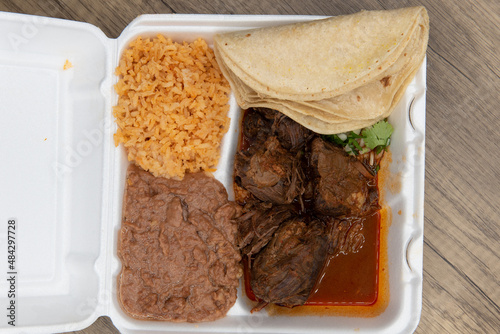 Overhead view of hearty take out order of birria stew meat served to make tacos with the included tortillas, rice and beans in the styrofoam box photo