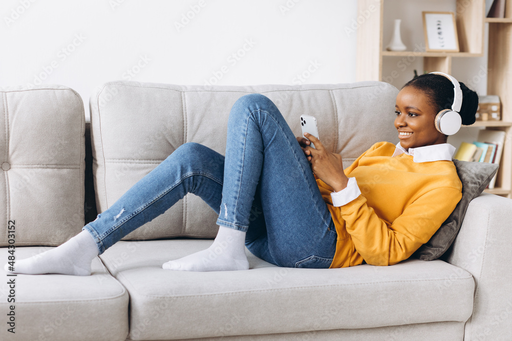 People, technology and leisure concept - happy african american young woman sitting on sofa with smartphone and headphones listening to music at home People, technology and leisure concept - happy african american young woman sitting on sofa with smartphone and headphones listening to music at home