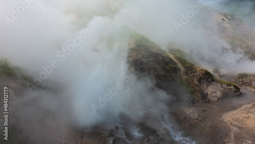 A geyser erupts on a mountainside. Splashes of boiling water are visible. Everything is shrouded in thick steam. The river flows along a rocky bed. Kamchatka. Valley of Geysers. Poor visibility.