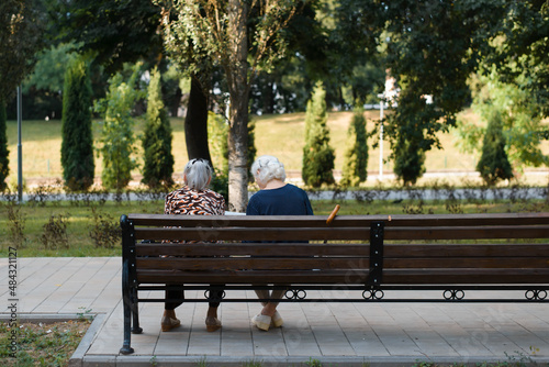 Back view of two old caucasian women girlfriends relaxing in park on summer day sitting on bench outdoors. Pensioners, elderly people leisure in nature
