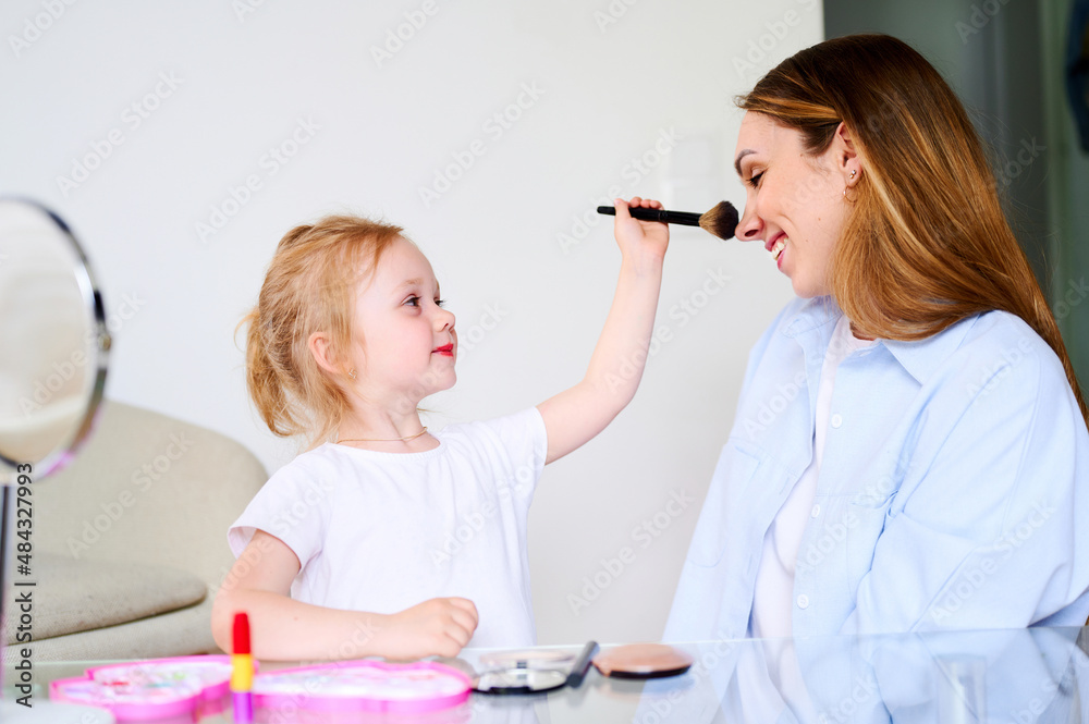 Mom and daughter play laugh and paint their lips with red lipstick. Mother teaches the child to do makeup. The relationship between parents and children Mom and daughter play laugh and paint their lips with red lipstick. Mother teaches the child to do makeup. The relationship between parents and children