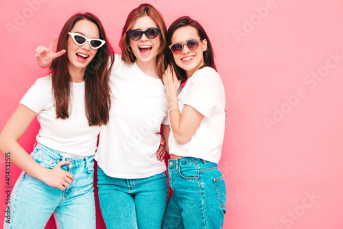 Three young beautiful smiling hipster female in trendy same summer white t-shirt and jeans clothes. Sexy carefree women posing near pink wall in studio. Positive models © halayalex