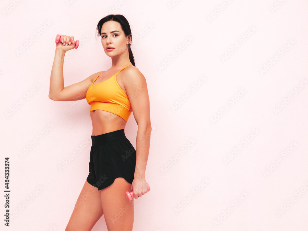 Fitness confident woman in black sports clothing. Sexy young beautiful model with perfect body. Female isolated on pink wall in studio. Stretching out before training.Making exercises with dumbbells Fitness confident woman in black sports clothing. Sexy young beautiful model with perfect body. Female isolated on pink wall in studio. Stretching out before training.Making exercises with dumbbells