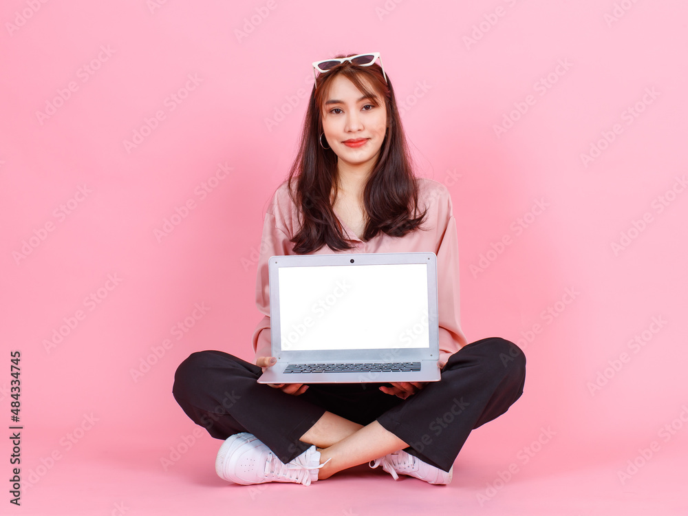 Naklejka premium Portrait studio shot of Asian young hipster female model in casual street wears sunglasses sit on floor holding pointing white blank empty screen laptop computer for advertising on pink background.