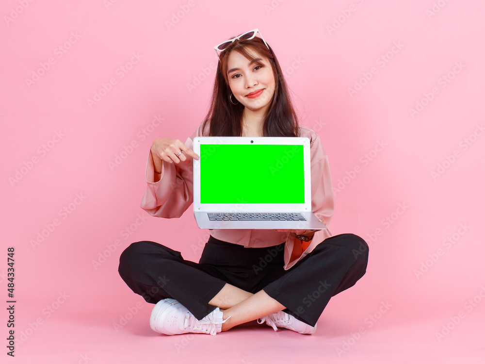 Portrait studio shot of Asian young hipster female model in casual street wears sunglasses sit on floor holding pointing green blank empty screen laptop computer for advertising on pink background Portrait studio shot of Asian young hipster female model in casual street wears sunglasses sit on floor holding pointing green blank empty screen laptop computer for advertising on pink background