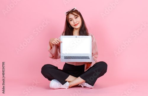 Portrait studio shot of Asian young hipster female model in casual street wears sunglasses sit on floor holding pointing white blank empty screen laptop computer for advertising on pink background.