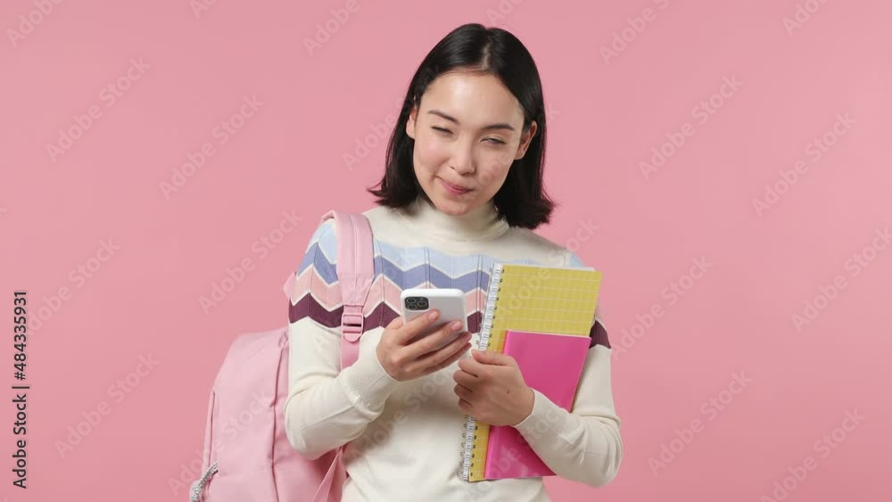 Minded girl teen student of Asian ethnicity wear shirt backpack hold books use mobile cell phone isolated on plain pastel light pink background studio portrait. Education in university college concept Minded girl teen student of Asian ethnicity wear shirt backpack hold books use mobile cell phone isolated on plain pastel light pink background studio portrait. Education in university college concept