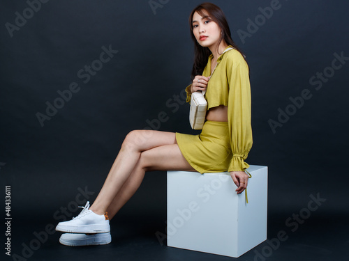 Portrait studio shot of Asian trendy modern fashion female model in casual crop top outfit sitting smiling on white box stool look at camera holding hands pointing fingers sideway on black background