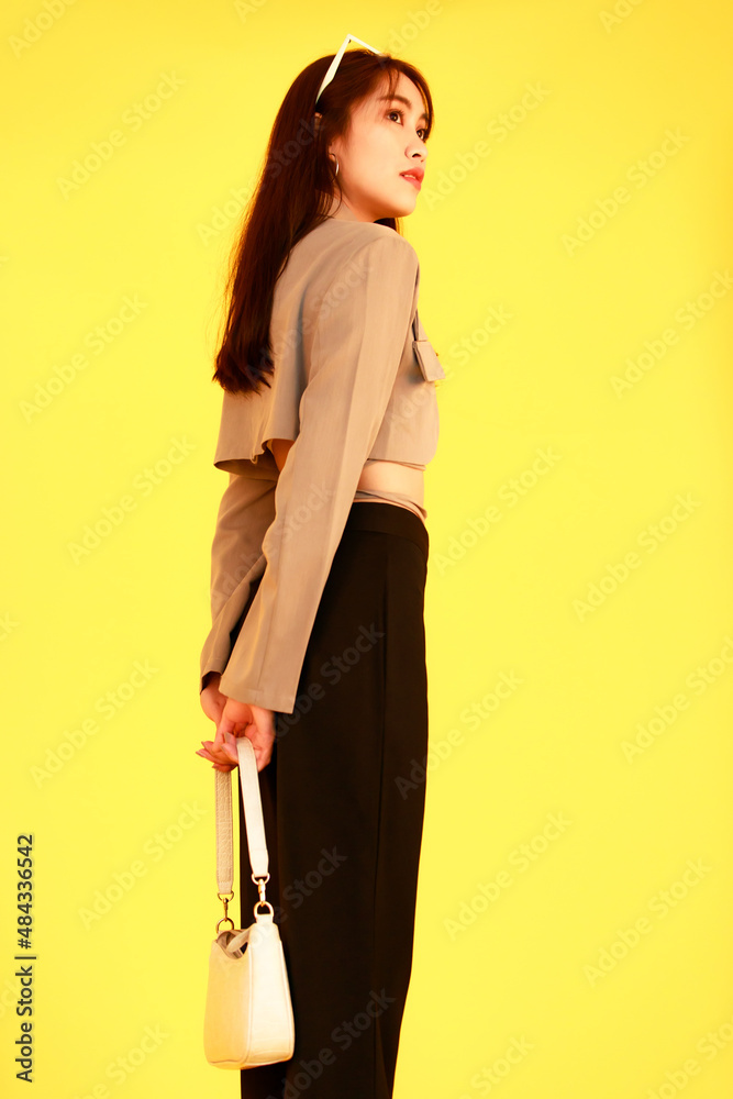 Portrait studio shot of Asian trendy fashionable female hipster teen model in casual crop top street wears jacket sunglasses sneakers holding handbag purse look at camera on yellow background