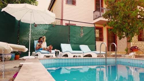 A male caucasian tourist lies on sunbed near the swimming pool in hotel and reading news in mobile in his hands. He relaxes surfing the internet on smartphone. Man frows then smiles watching at screen photo