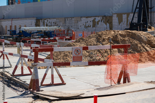 fenced area of the construction site. Red and white safety barriers enclose a pile of soil. There is warning sign on the barrier. Translation: 