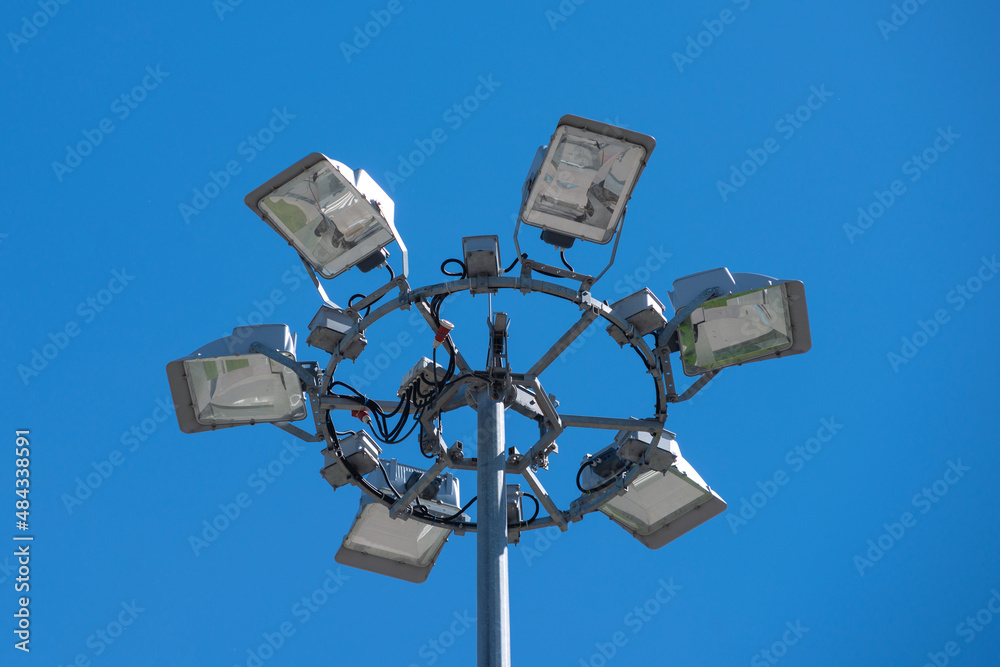 Projector stand made of several individual spotlights on the background of blue clear limitless sky. Lighting mast concept at the stadium, construction site or plant Projector stand made of several individual spotlights on the background of blue clear limitless sky. Lighting mast concept at the stadium, construction site or plant