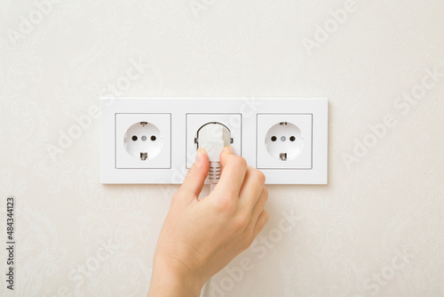 Young adult woman hand holding and plugging white electrical plug in wall outlet socket at home. Closeup. photo