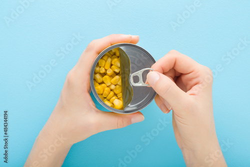 Принти на полотні Young adult woman hands holding and opening new metallic preserved can with yellow corn on light blue table background