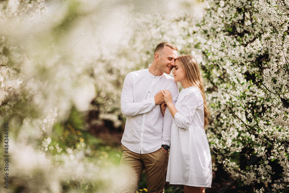 A happy young couple in love stands in a garden of blooming apple trees. A man in a white shirt and a girl in a white light dress are walking in a flowering park A happy young couple in love stands in a garden of blooming apple trees. A man in a white shirt and a girl in a white light dress are walking in a flowering park