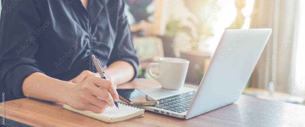 Woman hand writing on a notepad with a pen and use a laptop computer in the office.