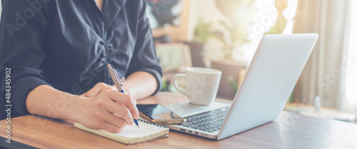 Woman hand writing on a notepad with a pen and use a laptop computer in the office.