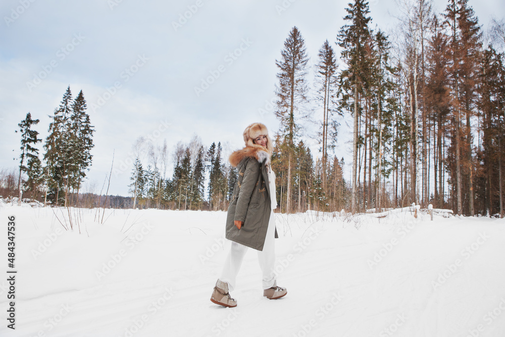 Obraz premium Happy smiling woman walking in winter snow nature meadow, hiking, wearing stylish fashion outfit khaki parka coat jacket, fur hat and moon boots, full length, looking back