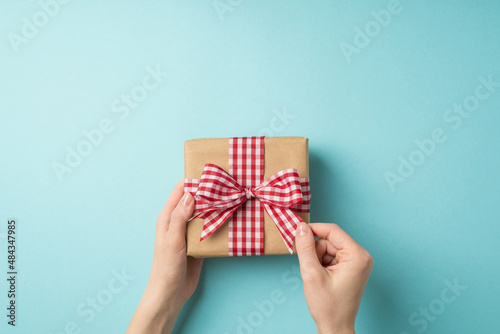 First person top view photo of valentine's day decorations girl's hands unpacking craft paper giftbox with checkered ribbon bow on isolated pastel blue background with copyspace