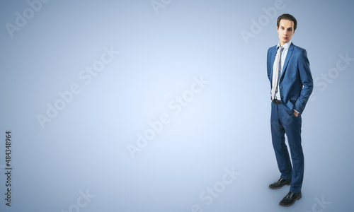 Job and recruiting concept with young businessman in blue classic suit, his hands in pockets on abstract light blue background with a space for your logo or text. Mockup