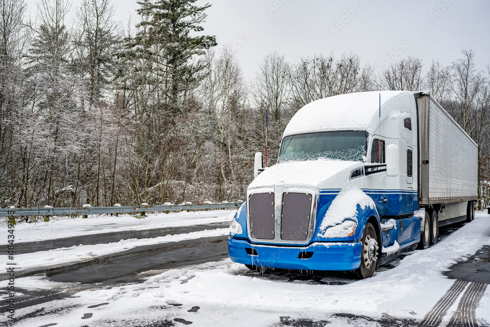 Professional long haul big rig semi truck with reefer semi trailer standing for rest on the winter truck stop parking lot with snow and ice and trees on the background