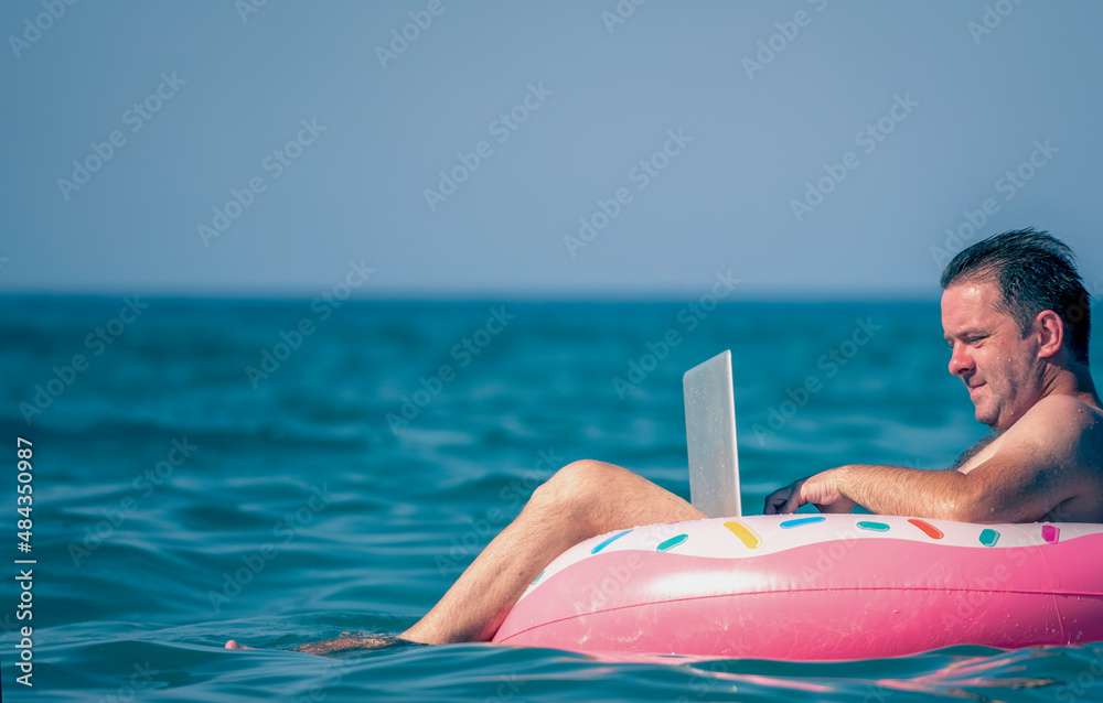 Working anywhere and any time concept. Young man working, using laptop computer on inflatable ring in the water. Freelance work, vacations, distance work, social distancing. Copy space for text
