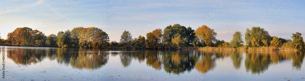 Panoramic photo of a pond and trees whose leaves are autumn colored. The surface of the ponds reflects the image of trees.