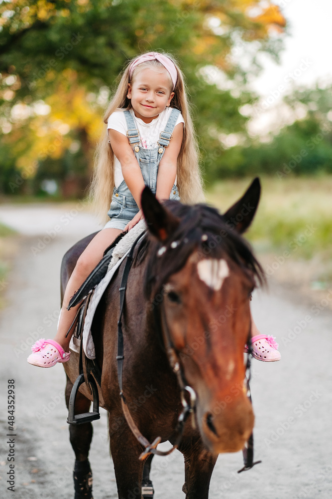 Fototapeta premium Little blonde girl with long hair rides a horse in the park at sunset in autumn. Autumn horse ride. Friendship of a girl and a horse.