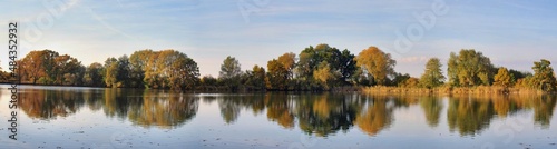 Panoramic photo of a pond and trees whose leaves are autumn colored. The surface of the ponds reflects the image of trees.