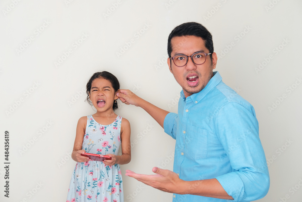 Asian father showing angry expression when tweak his daughter ear while holding mobile phone Asian father showing angry expression when tweak his daughter ear while holding mobile phone