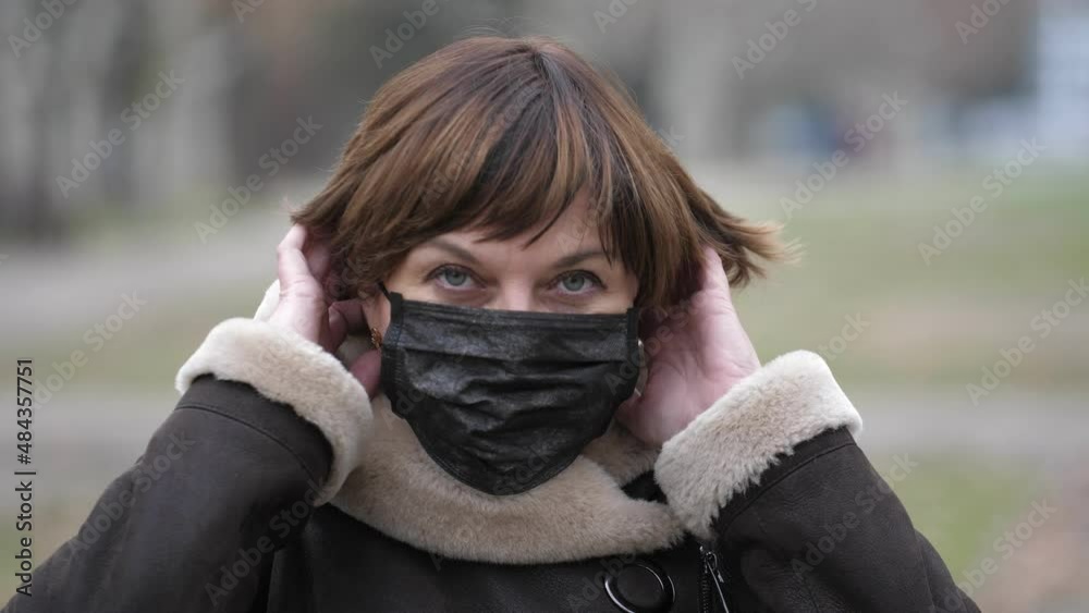 A beautiful European woman stands on the street, she looks into the camera and then puts on a black medical mask. Close-up, shallow depth of field A beautiful European woman stands on the street, she looks into the camera and then puts on a black medical mask. Close-up, shallow depth of field