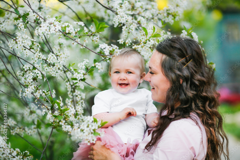 Lovely beautiful caucasian mom with her 1 year old daughter in spring blooming flowers. The concept of natural, conscious motherhood, female natural beauty and wavy hair.Young european woman with baby