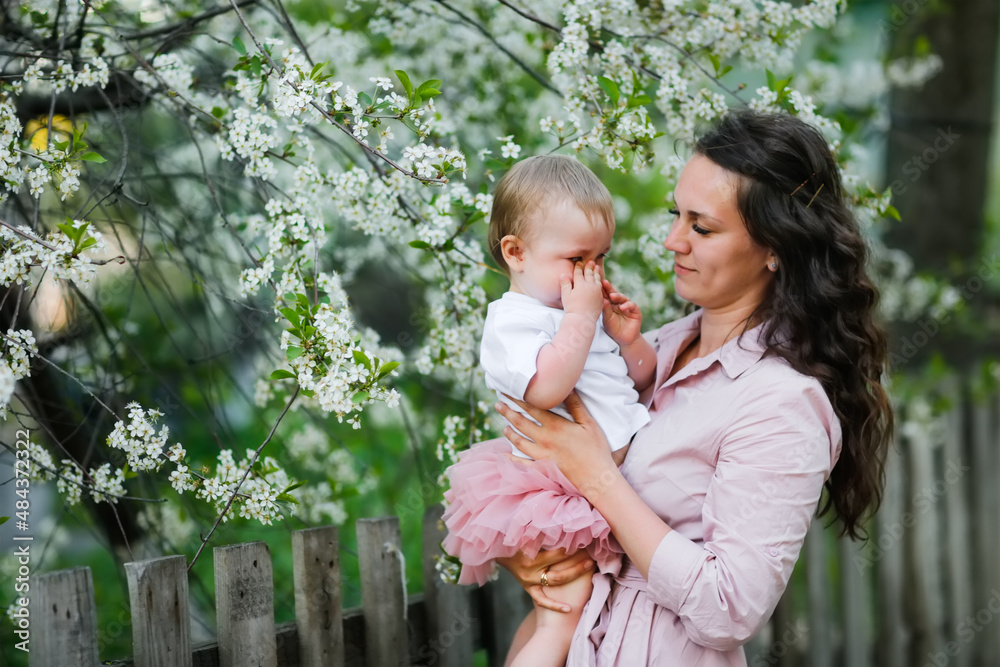 Fototapeta premium Lovely beautiful feminine mom with her daughter in her arms in spring with cherry blossoms. female natural beauty and beautiful wavy hair. Young European woman in a dress with a baby in a skirt.
