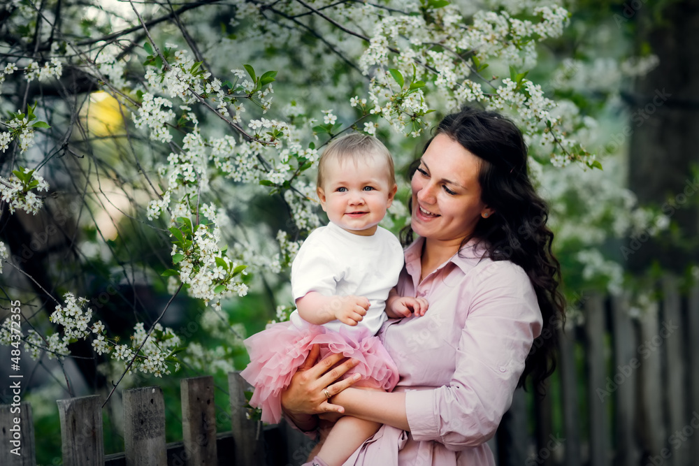 Lovely beautiful feminine mom with her daughter in her arms in spring with cherry blossoms. female natural beauty and beautiful wavy hair. Young European woman in a dress with a baby in a skirt.