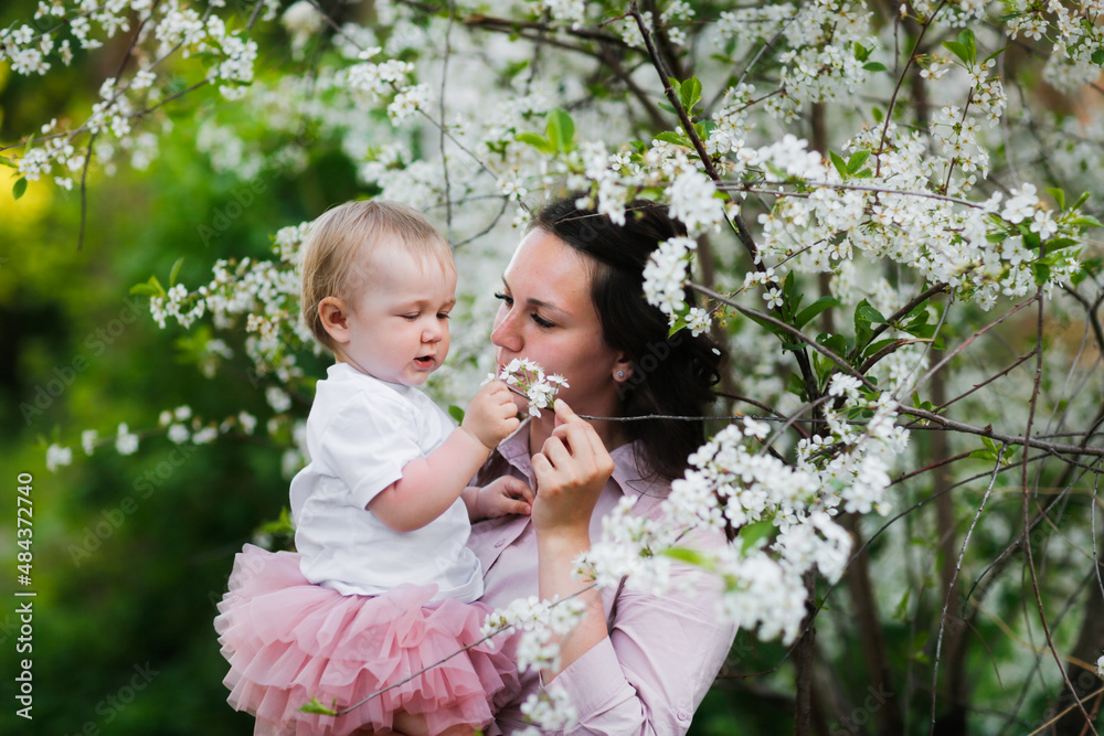 Obraz premium Lovely beautiful feminine mom with her daughter in her arms in spring with cherry blossoms. female natural beauty and beautiful wavy hair. Young European woman in a dress with a baby in a skirt.